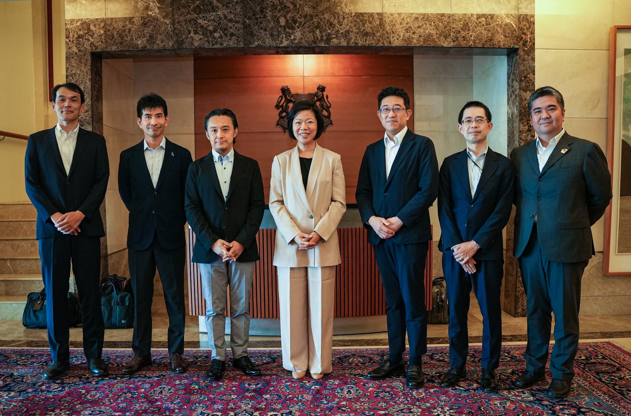 Seven people in suits stand in a row on a patterned rug.
